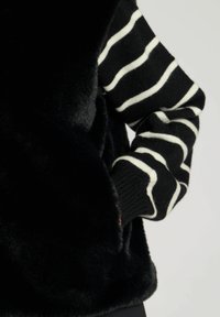 Svart fuskpälsväst med hög krage, kombinerad med en randig svart och vit långärmad tröja. Västen har en slät, mjuk textur.