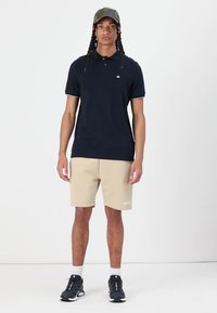 Navy polo shirt with short sleeves, beige shorts with white logo, and black athletic shoes. The model wears a green camo cap.