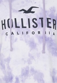 Fialová a bílá mikina s kapucí ve stylu tie-dye s nápisem "HOLLISTER CALIFORNIA" v tučném černém písmu a siluetou ptáka. Nastavitelná kapuce se šnůrkou.
