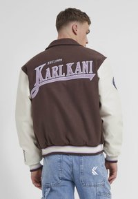 Hnedo-krémová varsity bunda s rebrovanými manžetami. Na zadnej strane má výšivku "KARL KANI" a dátum založenia, z materiálov texturovaná vlna a koža.