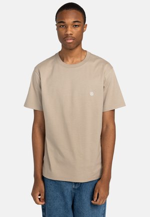 Beige katoenen t-shirt met een ronde hals en korte mouwen, voorzien van een klein logo op de linkerborst. Gedragen met blauwe denim shorts.