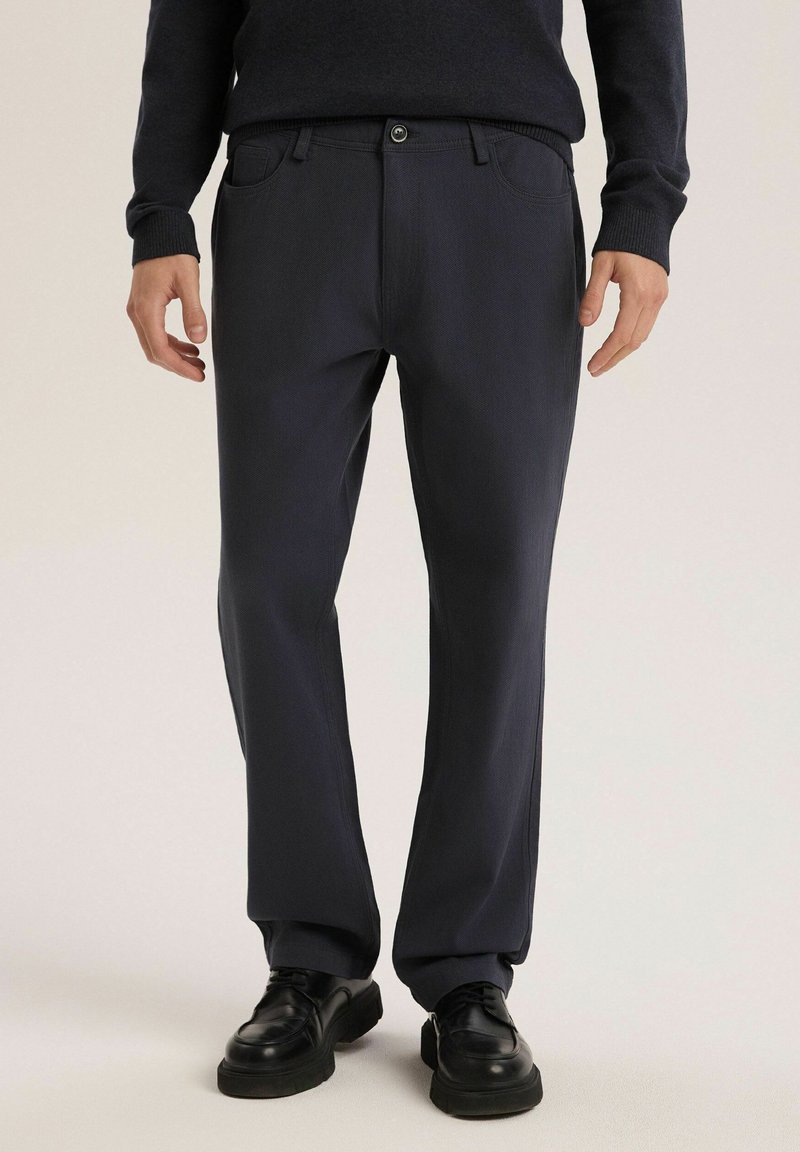 Granatowe spodnie wykonane z teksturowanej tkaniny, z zapięciem na guziki, bocznymi kieszeniami i luźnym krojem, zestawione z czarnymi butami.