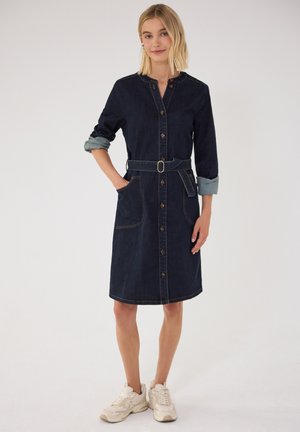 Femme portant une robe boutonnée en denim bleu foncé avec manches retroussées, taille ceinturée, poches avant et baskets blanches, debout devant un fond uni.