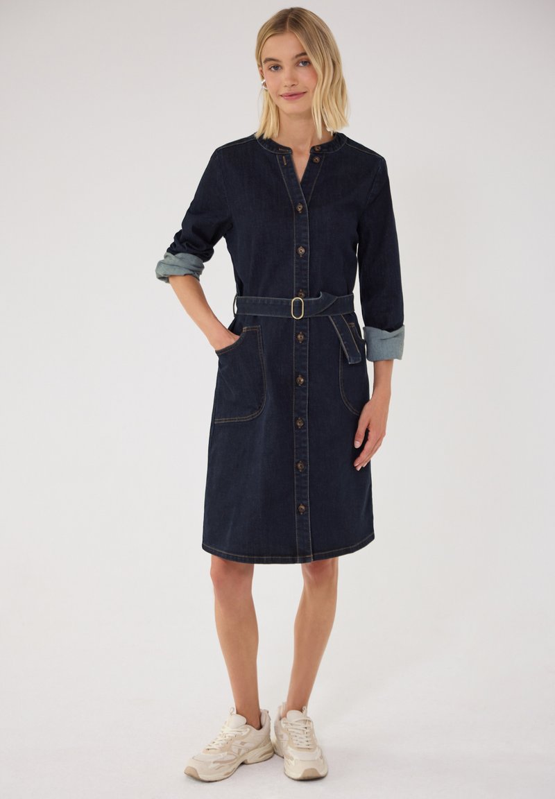 Femme portant une robe boutonnée en denim bleu foncé avec manches retroussées, taille ceinturée, poches avant et baskets blanches, debout devant un fond uni.