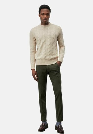 Maglione beige a trecce con collo rotondo, abbinato a pantaloni tailored verde scuro. Il modello indossa mocassini scuri e calzini blu.