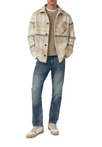 Beige karierte Flanelljacke mit Knopfschluss, kombiniert mit einem gestrickten hellbraunen Pullover und blauen Jeans. Weiße Sneaker mit hellbraunen Akzenten.