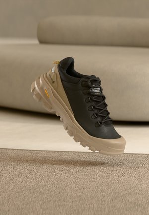 Zapato de senderismo negro y marrón con suela gruesa con dibujo, con marcas Gore-Tex y Vibram, fotografiado en el aire sobre una alfombra beige y un fondo de sofá.