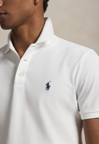 Prim-plan cu o persoană care poartă un tricou polo alb cu mâneci scurte, guler ridicat și un mic logo brodat bleumarin pe piept.