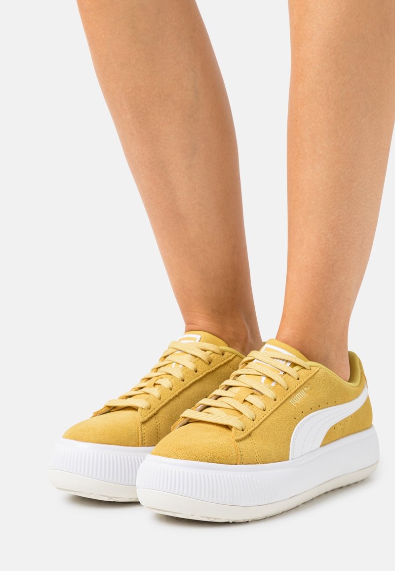 Yellow suede sneakers with white accents, rounded toe, and thick white rubber sole; features flat laces and logo detail on the side.