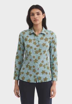 Frau trägt hellblaues Hemd mit grünen und orangefarbenen Blumenmustern und dunkle Hose, steht vor einfachem Hintergrund.