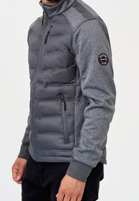 Mann trägt eine graue Jacke mit Reißverschluss, wattierter Vorderseite und strukturierten Ärmeln, mit einem runden Logo-Patch am Oberarm.