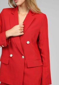 Femme ajustant le col d'un blazer rouge croisé avec des boutons argentés, se tenant devant un fond gris uni.