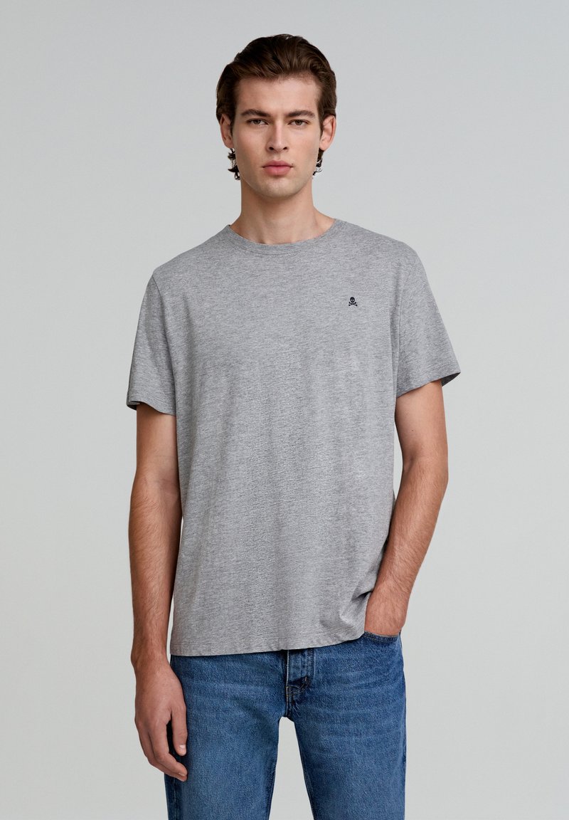 Grijze katoenen T-shirt met een ronde halslijn, korte mouwen en een klein zwart schedel logo op de borst, gecombineerd met blauwe jeans.
