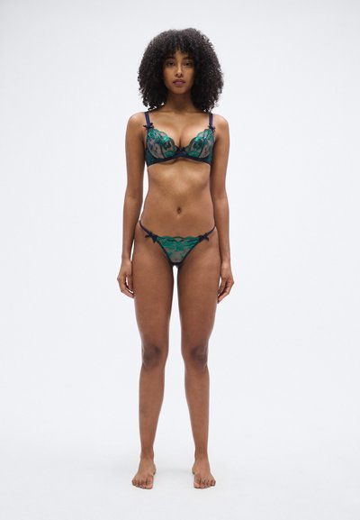 Ensemble de lingerie composé de dentelle verte à motif floral, bretelles bleu marine et nœuds. Le soutien-gorge est à armatures avec un design transparent ; le string vient le compléter.