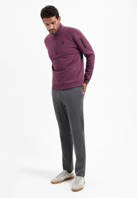 Pullover sweater van paarse stof met een kwartrits, voorzien van een zak met een zwarte ritssluiting en een geribde zoom. Gecombineerd met grijze, op maat gemaakte broek.