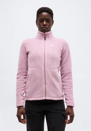 Person trägt eine hellrosa Fleecejacke mit Reißverschluss und Stehkragen sowie schwarze Hose und steht vor einem schlichten hellen Hintergrund.