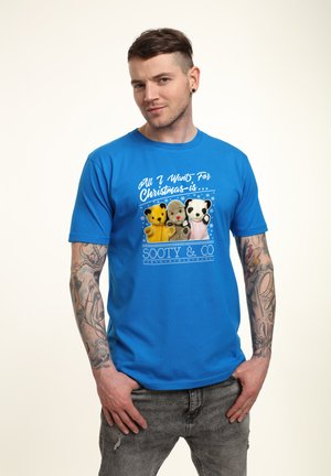 T-shirt en coton bleu avec un graphisme représentant quatre animaux en peluche, le texte indique "Tout ce que je veux pour Noël, c'est... Sooty & Co." Des flocons de neige décoratifs sont inclus.