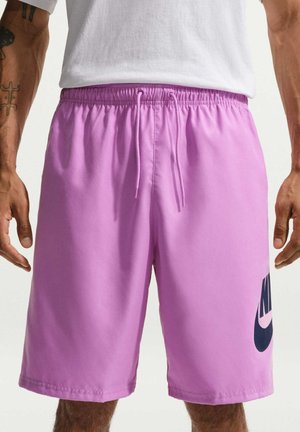 Mężczyzna noszący jaskrawo fioletowe spodenki Nike z paskiem do wiązania w talii i czarnym logo na prawej nodze, w połączeniu z białym T-shirtem.