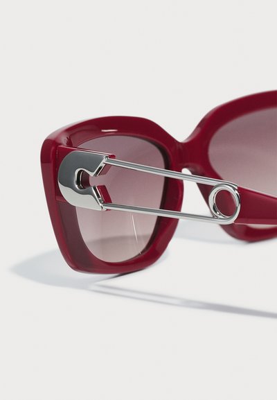 Óculos de sol de plástico vermelho com uma forma subtil de olho de gato, apresentando um detalhe em alfinete de segurança metálico de lado e lentes castanhas em degradê.