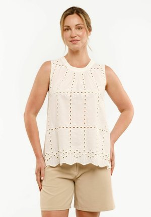 Kobieta ubrana w białą bezrękawnikową bluzkę z ażurowym wzorem oraz beżowe szorty do kolan, stojąca z jedną ręką na biodrze na białym tle.