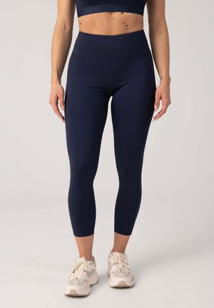 Femme portant des leggings courts taille haute bleu marine et des baskets blanches, debout devant un fond clair uni.