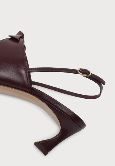 Κοντινή λήψη μπορντό slingback γόβας με λεπτό λουράκι στον αστράγαλο και μικρή χρυσή αγκράφα, σε απλό λευκό φόντο.