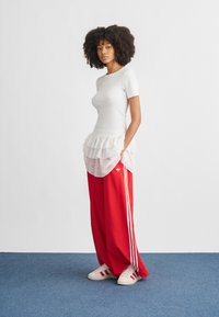 Žena s kudrnatými vlasy stojí na modrém koberci, má na sobě bílý přiléhavý top, vrstvenou bílou sukni přes červené Adidas tepláky a odpovídající tenisky.