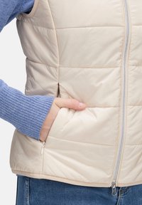 Beżowa kamizelka pikowana z pionowym wzorem, z zamkiem z przodu i kieszenią po bokach. Nosi ją osoba w jasnoniebieskim swetrze z prążkami.