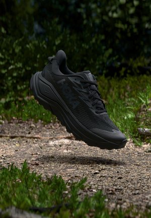 Melns Hoka skriešanas apavu pāris, kas karājas virs grants takas, pa perimetru apauguši ar zāli un krūmiem.