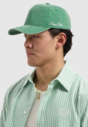 Man wearing a green "Pure Path" cap, light green striped button-up shirt with emblem, white undershirt, and gold necklaces, looking left against a plain background.