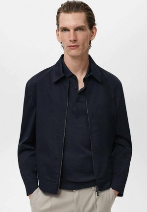 Marineblaues Zip-Jackett aus glattem Stoff, mit einem Kragen, Fronttaschen und einem lässigen Schnitt, kombiniert mit einem dunkelblauen Poloshirt darunter.