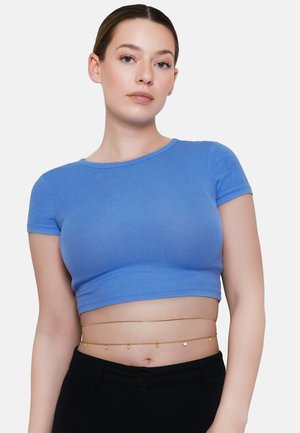 Junge Frau trägt ein blaues Cropped-T-Shirt und schwarze Hose, accessoriert mit einer filigranen goldenen Taillenkette mit kleinen Anhängern.