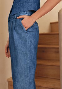 Mėlynos spalvos džinsinės kelnės laisvo stiliaus, su šoninėmis kišenėmis, lygiu paviršiumi ir subtiliu blizgesiu; derinamos su atitinkančiu viršumi.