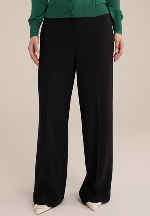 Femme portant un pantalon noir à jambes larges associé à des escarpins blancs pointus et un cardigan vert boutonné, posant devant un fond neutre.
