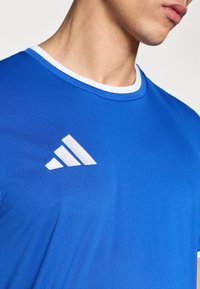 Camiseta deportiva azul con un cuello redondo blanco y un logo de tres rayas paralelas blancas en el pecho, confeccionada con un tejido ligero y suave.
