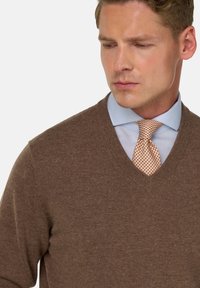 Rudas V formos megztinis su švelnia tekstūra, užsidėtas ant šviesiai mėlynos marškinėlių su apykakle, derinamas su raštuota oranžine kaklaraiščiu.