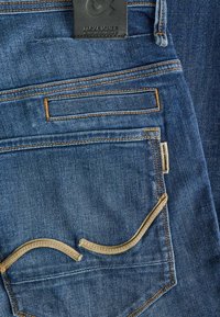 Blauwe denim jeans met gouden stiksels, zichtbare achterzakken, merknamen op zwart leren label en lichtbruine accenten op de rand van de zakken.