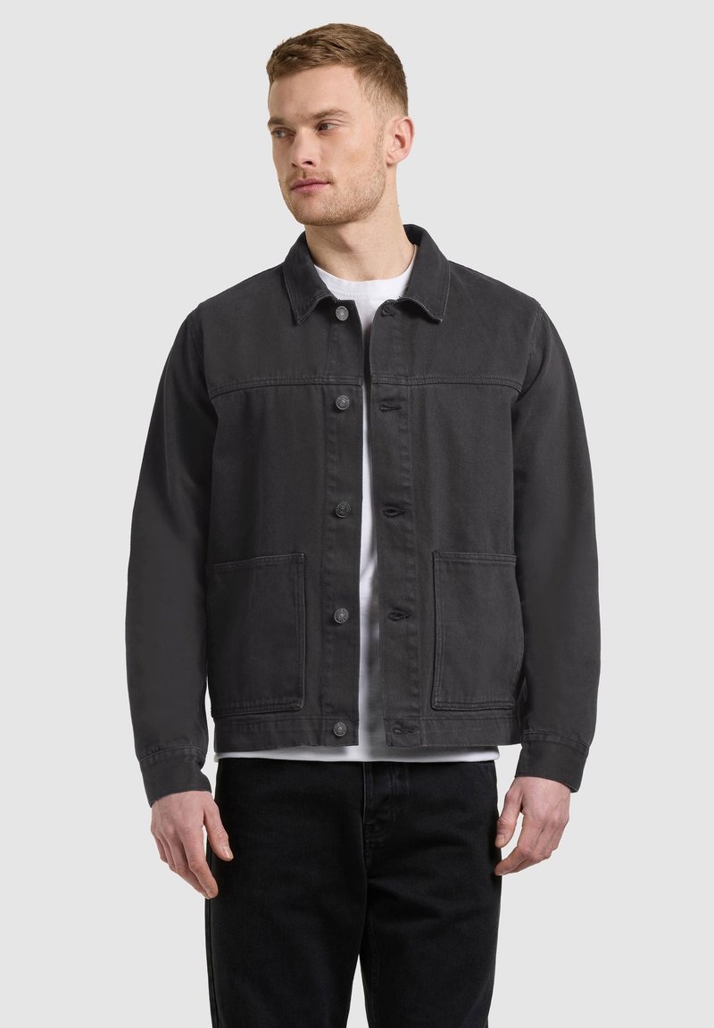 Homme portant une veste noire à boutons avec deux poches avant, par-dessus une chemise blanche, debout devant un fond clair uni.