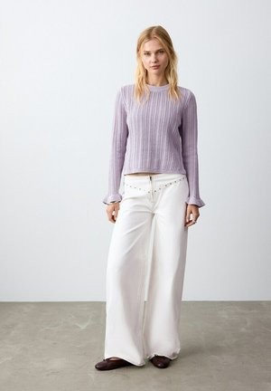 Donna con capelli biondi che indossa un maglione lavorato a maglia color lavanda, pantaloni bianchi a gamba larga con dettaglio di zip e scarpe piatte scure, in piedi su un pavimento in cemento.