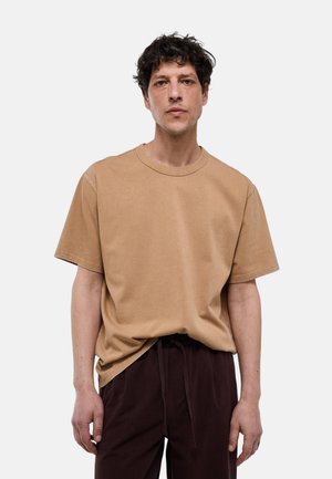 Homme aux cheveux bouclés foncés portant un t-shirt beige uni à manches courtes et un pantalon marron foncé à cordon, se tenant devant un fond blanc.