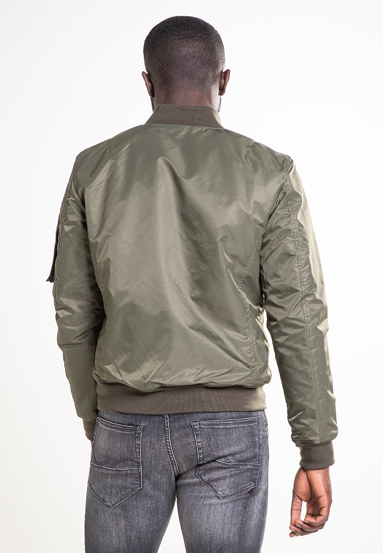 Homme aux cheveux coupés court portant une veste bomber olive et un jean gris, se tenant de dos face à l'appareil photo sur fond blanc.
