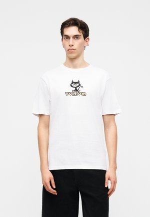 Νέος άνδρας με κοντά σκούρα μαλλιά φοράει λευκό μπλουζάκι Volcom με μικρό μαύρο καρτούν διάβολο και μαύρο παντελόνι, στέκεται μπροστά σε απλό φόντο.