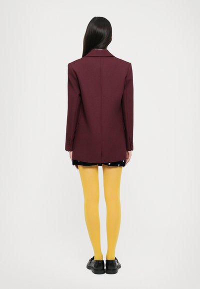 Woman standing facing away wearing a burgundy blazer, black floral skirt, yellow tights, and black shoes against a white background.