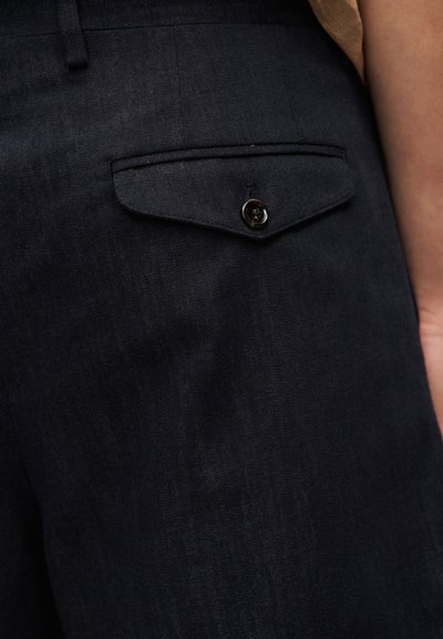 Κοντινό πλάνο σκούρου υφασμάτινου παντελονιού που δείχνει μια πίσω τσέπη με κλείσιμο με κουμπί και μέρος από το χέρι ενός ατόμου.