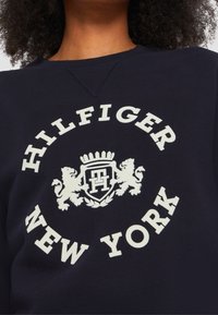 Marinblå tröja med centralt logotyp som har vit text "HILFIGER" och "NEW YORK", samt ett emblem med lejon och en krona.