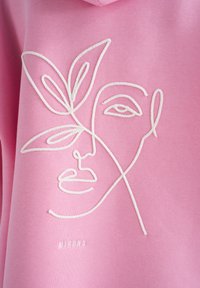 Felpa rosa con ricamo bianco di un viso e un design a foglia. Il tessuto ha una trama liscia con cuciture visibili e dettagli del logo.