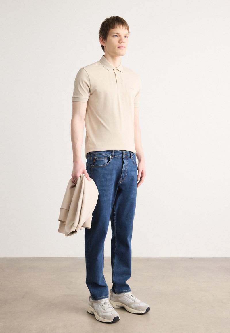 Polo beige à manches courtes, jeans en denim bleu et baskets grises. L'individu tient un sweat-shirt beige dans une main.