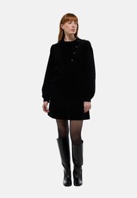 Robe en velours côtelé noir avec un décolleté carré, de longues manches bouffantes et des détails en boutons aux épaules, associée à des bottes noires jusqu'aux genoux.