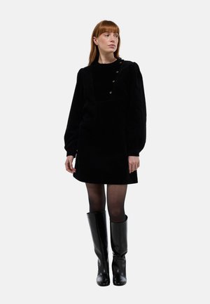 Robe en velours côtelé noir avec un décolleté carré, de longues manches bouffantes et des détails en boutons aux épaules, associée à des bottes noires jusqu'aux genoux.