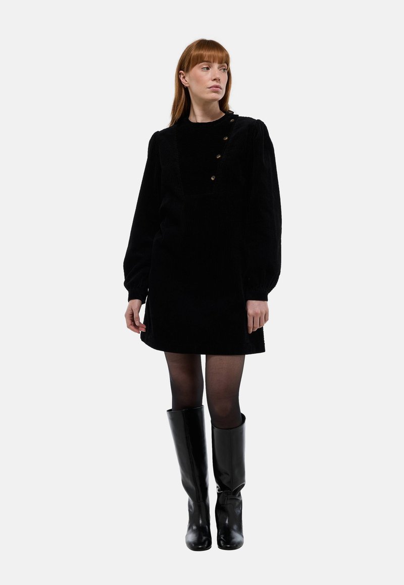 Robe en velours côtelé noir avec un décolleté carré, de longues manches bouffantes et des détails en boutons aux épaules, associée à des bottes noires jusqu'aux genoux.